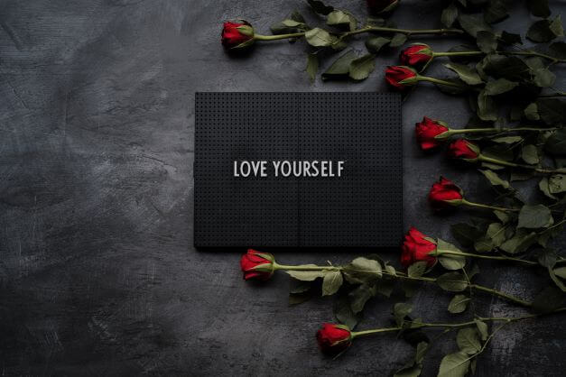 Love yourself written on a black sign with red roses nearby.