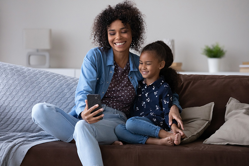 Mom on couch with smartphone and daughter smiling. Need a change? Therapy for moms in Detroit, MI may help. Call now and see how you could benefit from online therapy for new moms and any other service that may help!